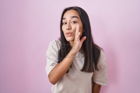Foto de Mujer hispana joven de pie sobre fondo rosa mano en boca contando rumores secretos, susurrando conversación maliciosa - Imagen libre de derechos