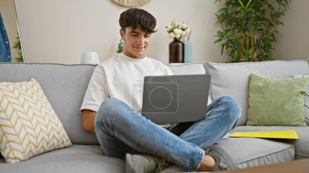 Photo for Handsome young hispanic teenager beams with joy, comfortably sitting on sofa, using laptop confidently at home, embodying happiness & relaxation in living room's warm interior. - Royalty Free Image