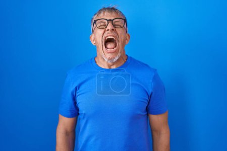 Foto de Hombre hispano con el pelo gris de pie sobre fondo azul enojado y loco gritando frustrado y furioso, gritando con ira. rabia y concepto agresivo. - Imagen libre de derechos