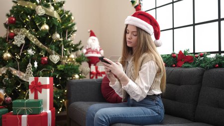 Foto de Mujer rubia joven usando teléfono inteligente sentado en el sofá por el árbol de Navidad en casa - Imagen libre de derechos
