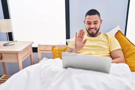 Foto de Hombre hispano usando portátil en la cama con aspecto positivo y feliz de pie y sonriendo con una sonrisa confiada mostrando los dientes - Imagen libre de derechos