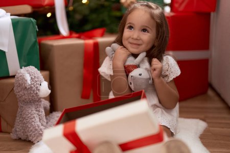 Photo for Adorable hispanic girl hugging teddy bear sitting on floor by christmas tree at home - Royalty Free Image