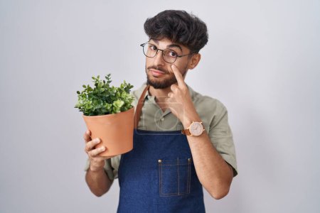 Téléchargez les photos : Arabe avec barbe tenant pot de plante verte pointant vers l'œil vous regardant geste, expression suspecte - en image libre de droit