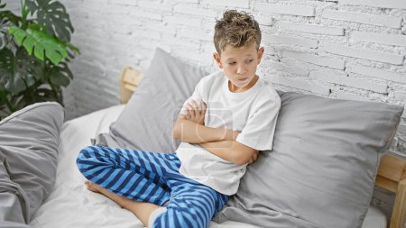 Adorable chico rubio, sentado en la cama con los brazos cruzados, mirando molesto en el dormitorio - un triste escenario de la mañana.