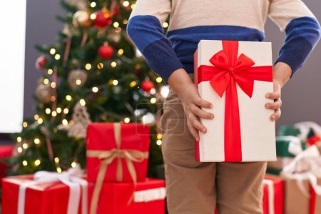 Foto de Adorable niño hispano sosteniendo regalo en la espalda de pie junto al árbol de Navidad en casa - Imagen libre de derechos