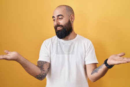 Téléchargez les photos : Jeune homme hispanique avec barbe et tatouages debout sur fond jaune souriant montrant les paumes ouvertes des deux mains, présentant et comparant la publicité et l'équilibre - en image libre de droit