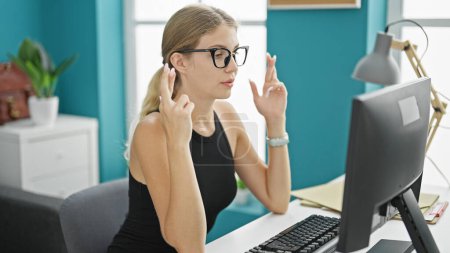 Foto de Joven mujer rubia trabajadora de negocios utilizando la computadora cruzando dedos para la suerte en la oficina - Imagen libre de derechos
