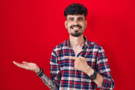 Téléchargez les photos : Jeune homme hispanique avec la barbe debout sur fond rouge étonné et souriant à la caméra tout en présentant avec la main et pointant du doigt. - en image libre de droit