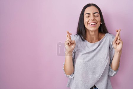 Téléchargez les photos : Jeune femme brune debout sur fond rose geste doigt croisé souriant avec espoir et les yeux fermés. chance et concept superstitieux. - en image libre de droit