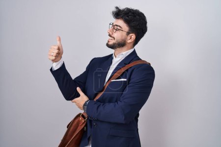 Téléchargez les photos : Homme hispanique avec barbe portant des vêtements de travail à l'air fier, souriant faire les pouces vers le haut geste sur le côté - en image libre de droit