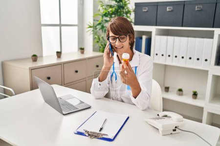 Photo for Young beautiful hispanic woman doctor talking on smartphone holding pills bottle at clinic - Royalty Free Image