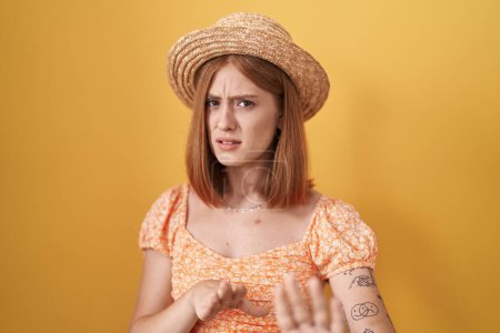Photo for Young redhead woman standing over yellow background wearing summer hat disgusted expression, displeased and fearful doing disgust face because aversion reaction. with hands raised - Royalty Free Image