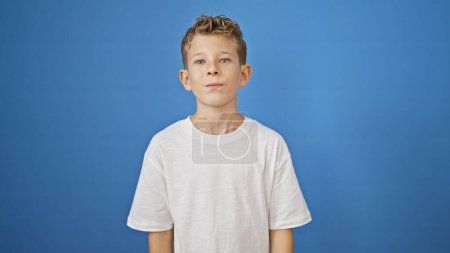 Foto de Adorable niño rubio de pie con una expresión seria en su cara, mirando atentamente a la cámara sobre un fondo azul aislado - Imagen libre de derechos