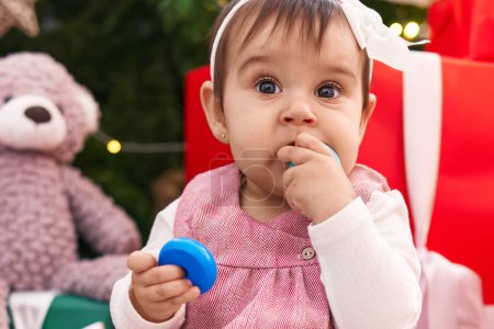 Foto de Adorable bebé hispano chupando el juguete del aro sentado en el suelo por el árbol de Navidad en casa - Imagen libre de derechos
