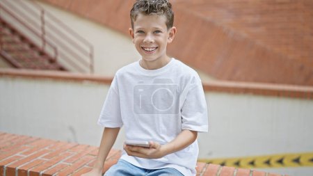 Photo for Adorable blond boy, cheerfully smiling while using a smartphone on a city street, engaged in a digital conversation, adding a touch of happiness to the urban landscape. - Royalty Free Image