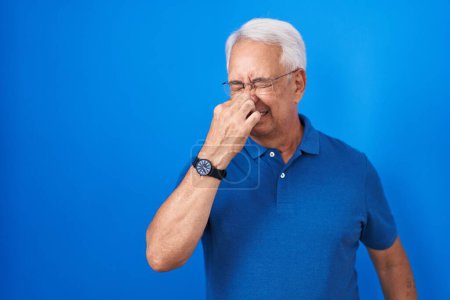 Téléchargez les photos : Homme d'âge moyen avec les cheveux gris debout sur fond bleu sentant quelque chose de puant et dégoûtant, odeur intolérable, retenant souffle avec les doigts sur le nez. mauvaise odeur - en image libre de droit
