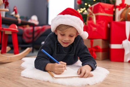 Foto de Adorable niño escribiendo en un cuaderno tumbado junto al árbol de Navidad en casa - Imagen libre de derechos