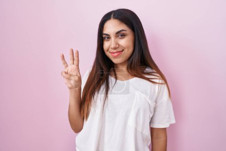 Téléchargez les photos : Jeune femme arabe debout sur fond rose montrant et pointant vers le haut avec les doigts numéro trois tout en souriant confiant et heureux. - en image libre de droit
