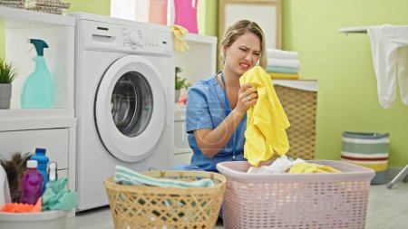 Téléchargez les photos : Jeune femme blonde nettoyeur professionnel sentant les vêtements sales à la buanderie - en image libre de droit