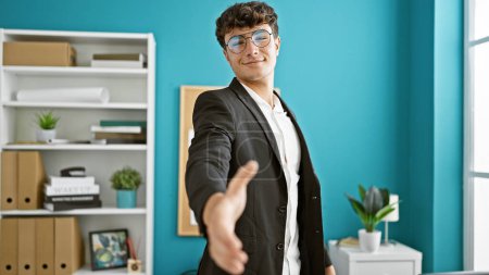 Téléchargez les photos : Jeune adolescent hispanique confiant travaille magie des affaires, souriant à la caméra tout en serrant fermement la main dans le bureau - en image libre de droit