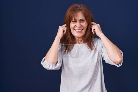 Foto de Mujer de mediana edad de pie sobre fondo azul cubriendo las orejas con los dedos con expresión molesta por el ruido de la música fuerte. concepto de sordo. - Imagen libre de derechos