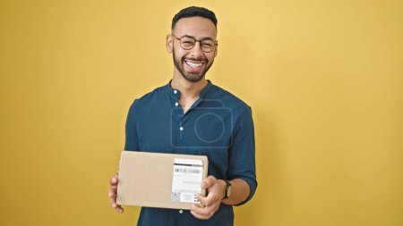 Téléchargez les photos : Jeune homme hispanique souriant tenant boîte en carton sur fond jaune isolé - en image libre de droit