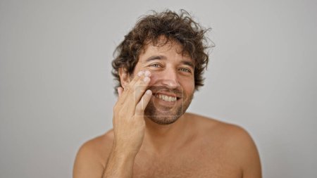 Téléchargez les photos : Jeune homme hispanique appliquant une lotion visage peau torse nu souriant sur fond blanc isolé - en image libre de droit