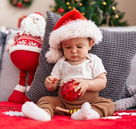 Téléchargez les photos : Adorable bébé hispanique tenant boule de décoration de Noël assis sur le canapé à la maison - en image libre de droit