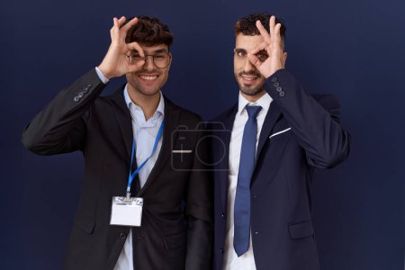 Téléchargez les photos : Deux hommes d'affaires hispaniques portant des vêtements de travail faisant un geste correct avec la main souriante, les yeux regardant à travers les doigts avec un visage heureux. - en image libre de droit