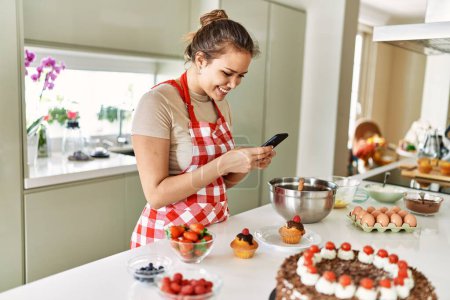 Téléchargez les photos : Jeune belle femme hispanique souriante confiante faire une photo par smartphone pour cupcake à la cuisine - en image libre de droit