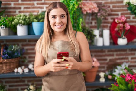 Téléchargez les photos : Jeune belle hispanique femme fleuriste souriant confiant en utilisant smartphone chez le fleuriste - en image libre de droit