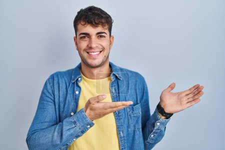 Foto de Joven hombre hispano de pie sobre fondo azul invitando a entrar sonriendo natural con la mano abierta - Imagen libre de derechos