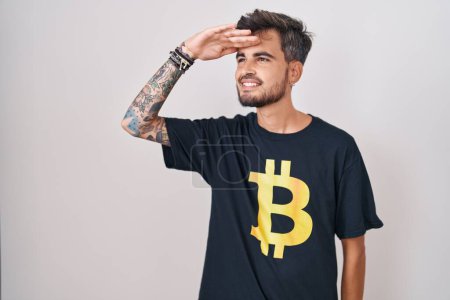 Photo for Young hispanic man with tattoos wearing bitcoin t shirt very happy and smiling looking far away with hand over head. searching concept. - Royalty Free Image