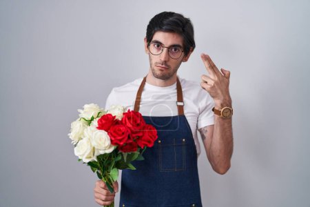 Foto de Joven hispano sosteniendo ramo de rosas blancas y rojas disparando y matándose apuntando de la mano y los dedos a la cabeza como arma, gesto suicida. - Imagen libre de derechos