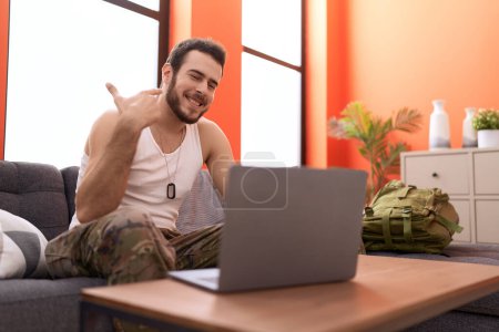 Téléchargez les photos : Jeune homme hispanique portant un uniforme de camouflage de l'armée faisant appel vidéo à la maison souriant heureux pointant avec la main et le doigt - en image libre de droit