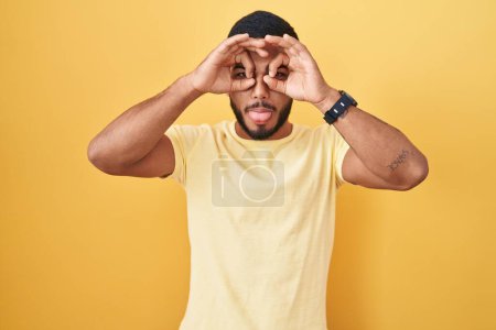 Photo for Young hispanic man standing over yellow background doing ok gesture like binoculars sticking tongue out, eyes looking through fingers. crazy expression. - Royalty Free Image