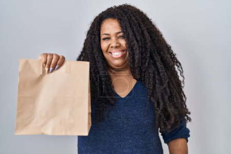 Foto de Más tamaño mujer hispana sosteniendo llevar bolsa de papel mirando positiva y feliz de pie y sonriendo con una sonrisa confiada mostrando los dientes - Imagen libre de derechos