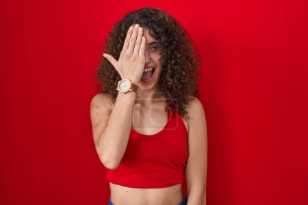 Foto de Mujer hispana con el pelo rizado de pie sobre fondo rojo cubriendo un ojo con la mano, sonrisa confiada en la cara y emoción sorpresa. - Imagen libre de derechos