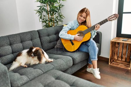 Téléchargez les photos : Jeune femme caucasienne jouant de la guitare classique assise sur un canapé avec chien à la maison - en image libre de droit