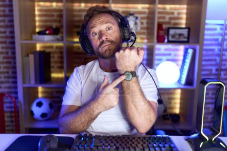 Téléchargez les photos : Homme du Moyen Age avec barbe jouant à des jeux vidéo portant des écouteurs pressés pointant vers le temps de regarder, impatience, regardant la caméra avec une expression détendue - en image libre de droit