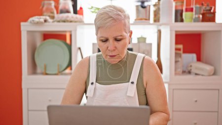 Photo for Middle age grey-haired woman using laptop sitting on table with serious face at dinning room - Royalty Free Image
