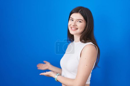 Téléchargez les photos : Jeune femme caucasienne debout sur fond bleu invitant à entrer souriant naturel avec la main ouverte - en image libre de droit