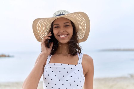 Photo for Young african american woman wearing summer hat talking on smartphone at seaside - Royalty Free Image