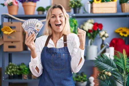Téléchargez les photos : Jeune femme blonde travaillant au magasin de fleuriste tenant des dollars criant fiers, célébrant la victoire et le succès très excité avec les bras levés - en image libre de droit