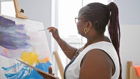 Foto de Artista afroamericana enfocada, con trenzas y gafas, dibujando seriamente sobre lienzo en estudio de arte interior - Imagen libre de derechos