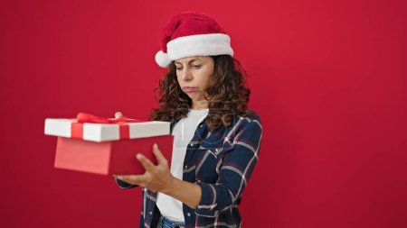 Téléchargez les photos : Moyen Age femme hispanique portant chapeau de Noël déballage cadeau à la recherche bouleversé sur fond rouge isolé - en image libre de droit