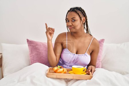 Téléchargez les photos : Femme afro-américaine avec des tresses tenant plateau avec de la nourriture pour le petit déjeuner dans le lit souriant heureux pointant avec la main et le doigt sur le côté - en image libre de droit