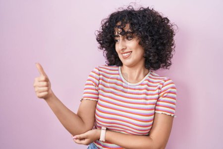 Photo for Young middle east woman standing over pink background looking proud, smiling doing thumbs up gesture to the side - Royalty Free Image