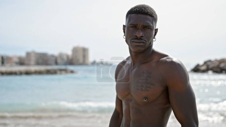Foto de Hombre africano americano turista de pie sin camisa con cara seria en la playa - Imagen libre de derechos