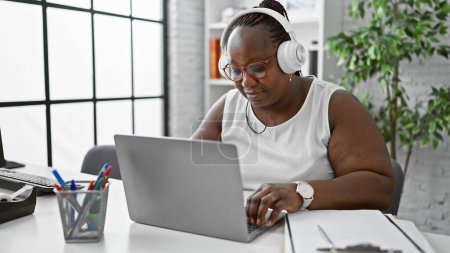 Foto de Prospera mujer afroamericana jefe trabajando duro en el ordenador portátil, el uso de auriculares en la oficina de negocios, exudando éxito profesional - Imagen libre de derechos